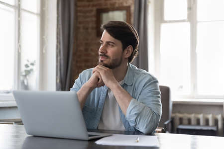 Thoughtful dreamy business professional man working at laptop in home office, looking at window, thinking, smiling at project future vision. Happy millennial freelancer in casual candid portraitの写真素材