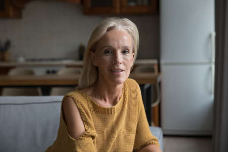 Positive mature lady home head shot portrait. Blonde middle aged pensioner, homeowner, retired woman in casual sitting on couch in living room, looking at camera, smilingの写真素材