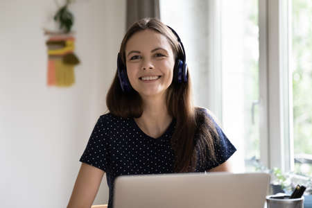 Woman in wireless headphones sit at desk with laptop staring at camera smile feels happy enjoy easy,...