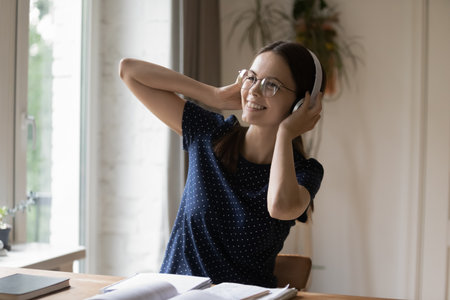 Happy woman listen song through wireless headphones sit at desk with textbook, take break from study, enjoy quality sound, favourite track at home use modern tech, digital stream music service conceptの写真素材
