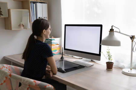 Rear view young woman sit at workplace desk with digital graphic tablet, staring at computer white mock up screen looks interested working or learns professional photo or video editor program conceptの写真素材