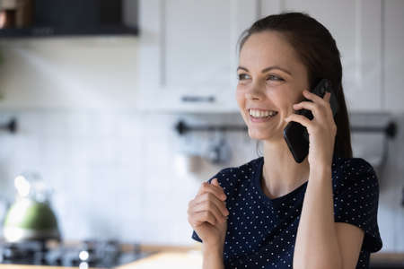 Cheerful young woman standing in kitchen holds smartphone having conversation, arrange meeting, talks with friend. Mobile operator usage, connection, communication, make food order delivery conceptの写真素材