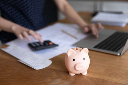 Unknown woman pay bills via ebank app on laptop, use calculator counts expenses, manage finances, busy in personal money and budget control, close up focus piggybank on table, economy, savings conceptの写真素材