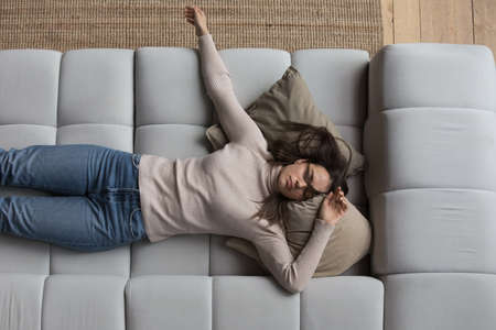 Sleepy millennial girl in casual clothes resting on couch at home, with spread hands and hair, sleeping at daytime, feeling tired, enjoying relaxation, leisure, recovery, reloading mind. Top viewの写真素材