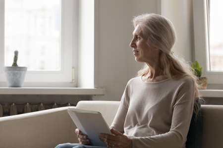 Pensive distracted 65s woman sit on sofa with digital tablet staring aside look thoughtful deep lost...