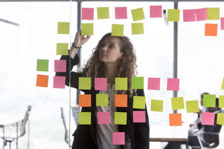 Focused young office employee working on project, using glass board, colorful paper tickers, writing...