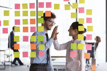 Happy millennial diverse business team friends giving high five, celebrating success at colorful notes on glass boards. Employees satisfied with scrum management work resultの写真素材