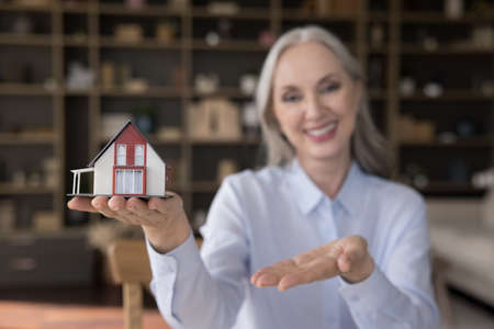Happy senior agent, realtor woman, seller showing tiny house model at camera, offering apartment for...