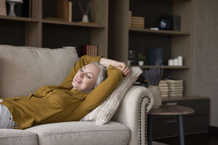 Carefree happy retired woman enjoying break, leisure time at home, relaxing, lying on comfortable couch, smiling, laughing, looking away, breathing fresh air. Stress relief conceptの写真素材