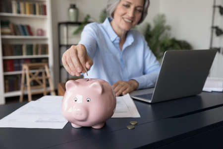 Happy senior retired lady making savings, financial reserve, putting money into pink ceramic piggybank. Tenant, homeowner, business woman counting personal budget, using piggy bankの写真素材