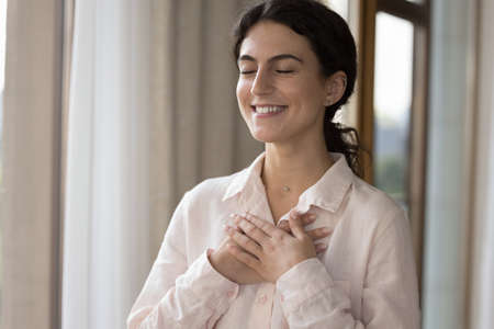 Young lovely woman standing alone indoors closed her eyes put folded palms on chest looks grateful,...