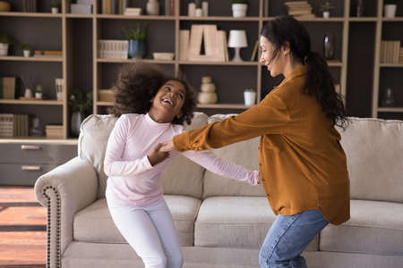 Cute little girl and mom hold hands dancing in living room. Young woman preschool daughter moving to favourite music looking carefree, feel overjoyed. Family enjoy hobby, spend leisure at home conceptの写真素材