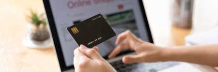 Hand of young woman holding black plastic credit card with copy space close up. Shopper, bank customer girl using online financial app on laptop computer, making paymentsの写真素材