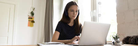 Focused millennial student girl in wireless headphones watching learning video tutorials, webinar,...