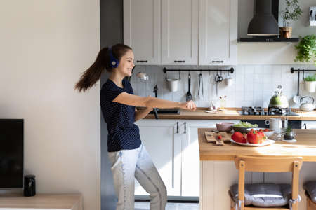 Cheerful active teen girl in earphones dancing in kitchen, enjoying cooking party, home entertainment, having fun, singing songs at ladle, cooking salad for healthy dinner at table with vegetableの写真素材