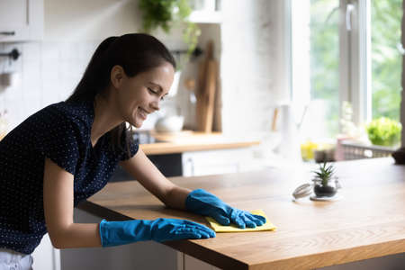 Happy tenant girl in blue protective rubber gloves enjoying cleanup at home, washing tabletop...