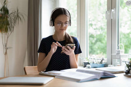 Focused smart student girl in wireless headphones using learning app on mobile phones, watching webinar on smartphone, listening to audio course, studying at open book, copy book at homeの写真素材