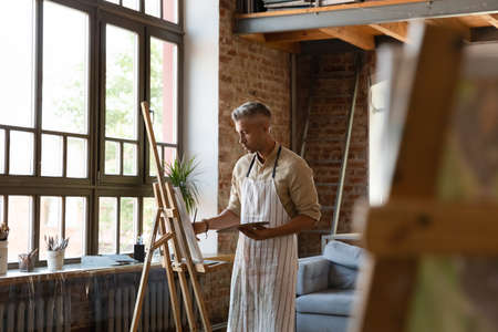 Serious man painter holds palette and paintbrush drawing on canvas in spacious loft workshop room. Talented male artist creates masterpiece, creative hobby, professional occupation, vocation conceptの写真素材
