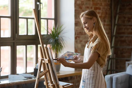 Young beautiful painter woman standing alone in light modern workshop painting picture on canvas with paints looks absorbed, smiles enjoy creative hobby for soul. Education, talent, art-class conceptの写真素材