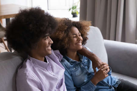 Happy excited couple of Black teenagers having fun, laughing, hugging, resting in couch. Cheerful...