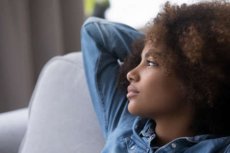 Pensive teenage African girl with natural curly hair resting on home couch, looking away in deep...