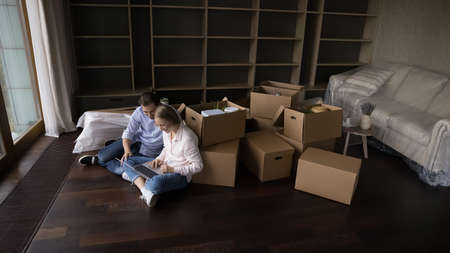 Top view in unfurnished living room couple use laptop sit on floor, heap of boxes with stuff nearby. Move day, e-services for comfort life, e-commerce clients choose buy furniture for new home conceptの写真素材