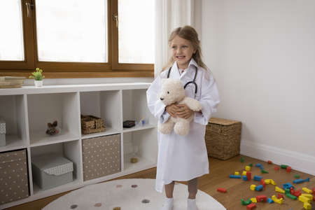 Little cute girl in medical coat and stethoscope play hospital at home, imagine herself veterinarian or nurse cure best fluffy friend. Paediatrician, healthcare, professional medicare services conceptの写真素材