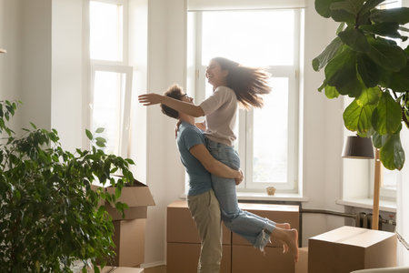 Happy excited young couple celebrating relocation, buying flat, apartment, moving in together, dancing, having fun, laughing. Guy holding girlfriend in arms, spinning at cardboard boxesの写真素材
