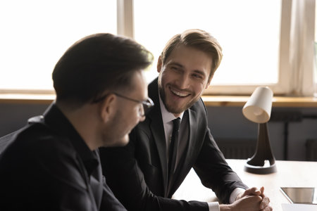 Happy young handsome businessman discussing work project with colleagues, enjoying cooperation, partnership, business success, sharing successful ideas. Two coworkers meeting at office tableの写真素材