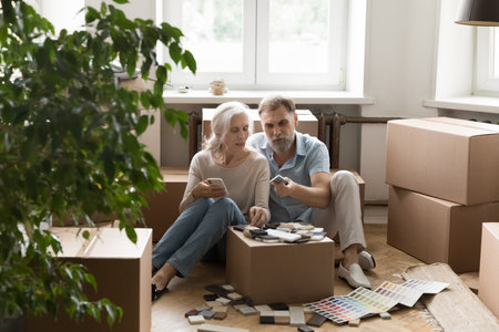 Elder grey haired family couple of new homeowners reviewing floor surface samples, choosing decoration colors, planning renovation, discussing interior design, sitting on floor at cardboard boxesの写真素材