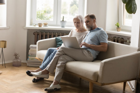 Engaged retired grey haired couple using laptop computer on couch together, chatting online, enjoying leisure, internet communication together, shopping, browsing social media. Full lengthの写真素材