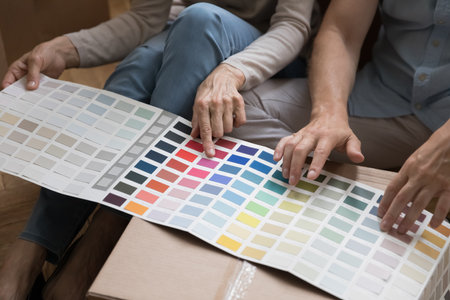 Senior couple discussing home renovation, pointing fingers at color swatches in paper brochure on cardboard box, choosing colors for new interior design. Cropped shot, close up of handsの写真素材