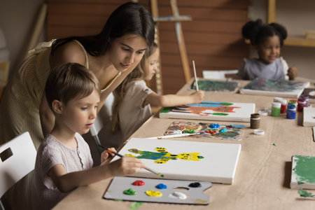 Friendly young art teacher giving artistic lesson to kids, helping to draw colorful cartoon animals, blend colors on palette, painting on canvas. Diverse group of children training creativityの写真素材