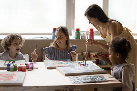 Happy pretty art school teacher giving lesson to pupil kids, bending over table with canvas, paints, palettes in creative studio, speaking, helping children to mix colors, draw artworkの写真素材