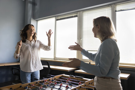 Two cheerful positive diverse business colleagues women playing table soccer, enjoying board game,...