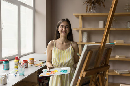 Happy beautiful young art school student drawing on canvas at easel, using artistic supplies, holding palette with acrylic, oil paints, looking at camera, smiling. Craft professional portraitの写真素材