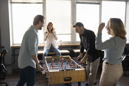 Happy joyful team of business colleagues having fun in office. Employees giving support to coworkers playing table soccer, tabletop football, laughing, smiling, shoutingの写真素材