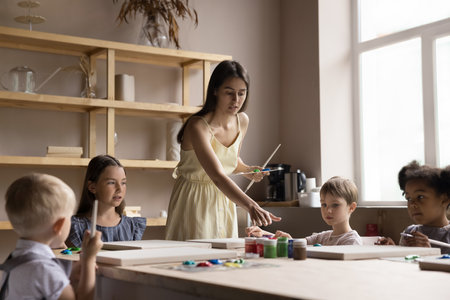 Pretty young artist woman teaching diverse group of little kids in artistic school studio. Children drawing pictures on class in acrylic, oil paints with assistance of female teacherの写真素材