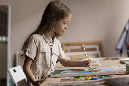 Focused little pupil girl drawing on canvas at table in classroom, training artistic creative...