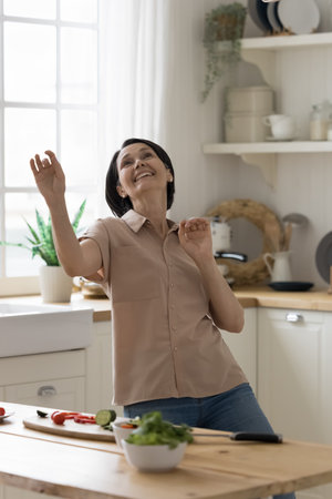Happy energetic mature lady having fun at home, cooking salad in kitchen, preparing healthy meal, keeping raw food eating, organic vegetarian diet, dancing at table, laughingの写真素材