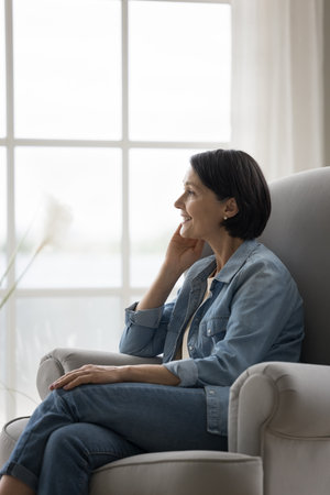 Happy dreamy pretty mature woman relaxing in armchair at home, thinking on good future plans, touching chin, looking away, smiling, dreaming. Positive senior lady enjoying leisure at homeの写真素材