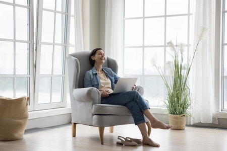 Cheerful middle aged woman using laptop at home, relaxing in armchair with gadget, talking on video call, watching funny movie online, laughing out loud, having fun, enjoying communication, leisureの写真素材