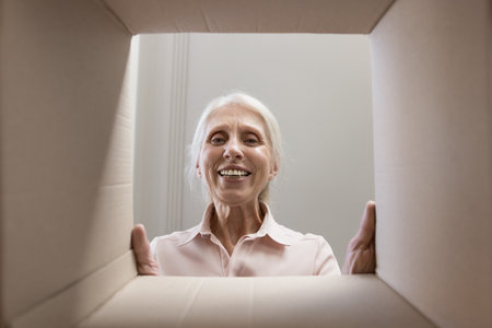 Happy joyful senior woman looking at camera through cardboard box dimensions frame, smiling, laughing. Elder customer lady checking paper container. From inside view portraitの写真素材