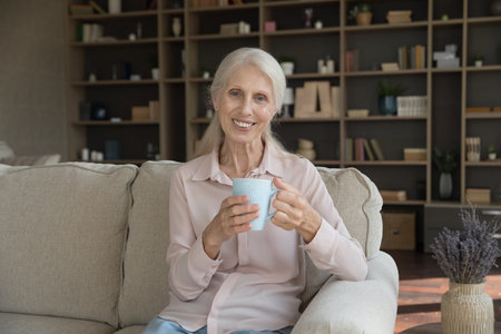 Positive older senior woman drinking hot coffee, tea at home, enjoying good morning, sitting on...