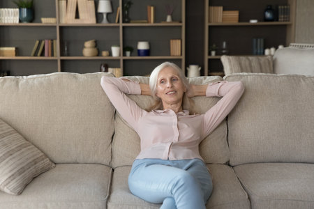 Thoughtful positive old senior woman resting on comfortable sofa, stretching body, breathing fresh cool air at home, leaning on couch back, dreaming, smiling, enjoying relaxation, leisureの写真素材