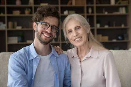 Cheerful handsome adult son and senior mom sitting close, looking at camera with toothy smiles, posing for shooting at home, talking on video call. Screen view, head shot portraitの写真素材