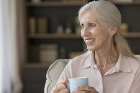 Positive older senior woman drinking hot coffee, tea at home, enjoying good morning, sitting on...