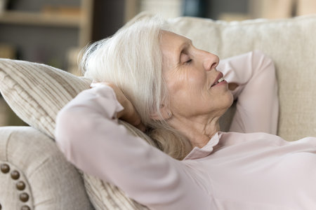 Relaxed sleepy older woman resting on comfortable couch, sleeping, taking nap, enjoying leisure, relaxation at cozy home, breathing fresh cool air, smiling, dreamingの写真素材