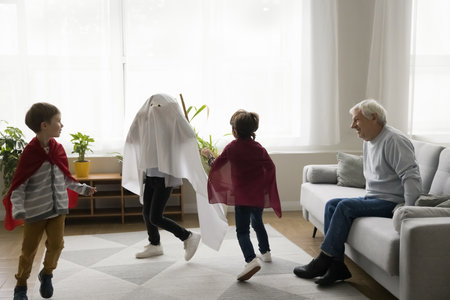 Happy senior grandfather watching three boys in homemade super hero costumes chasing each other, running, playing role active game. Grandpa enjoying meeting with grandsonsの写真素材