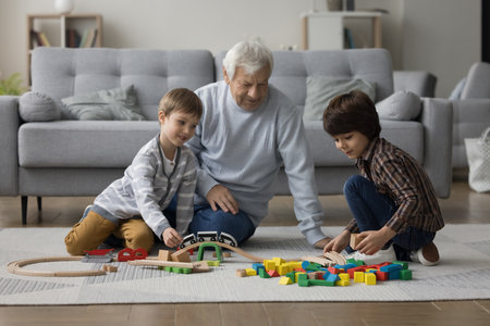 Two cheerful kids and grandpa playing constructor game on heating carpeted floor at home, building city road from mess of toy blocks. Grandkids visiting grandpa, having fun, enjoying leisureの写真素材